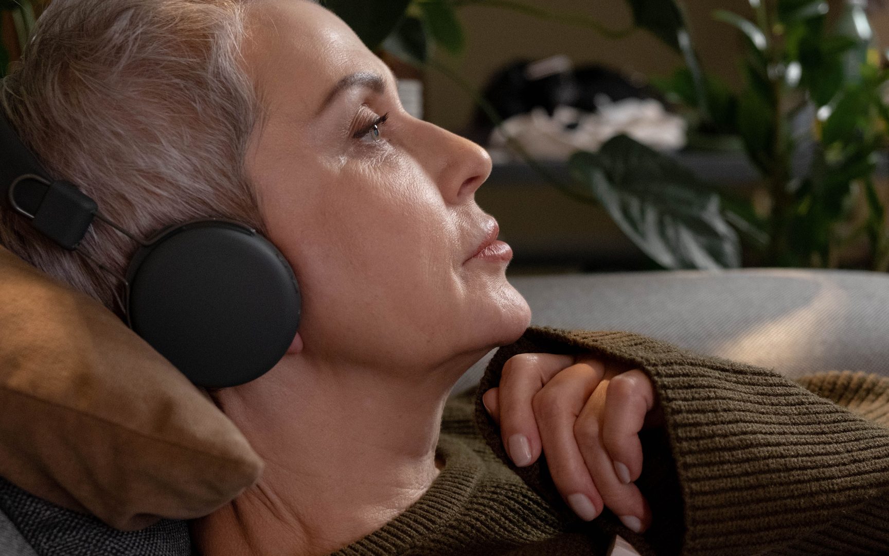 A woman resting back on a sofa wearing over-ear headphones, eyes open and gaze soft