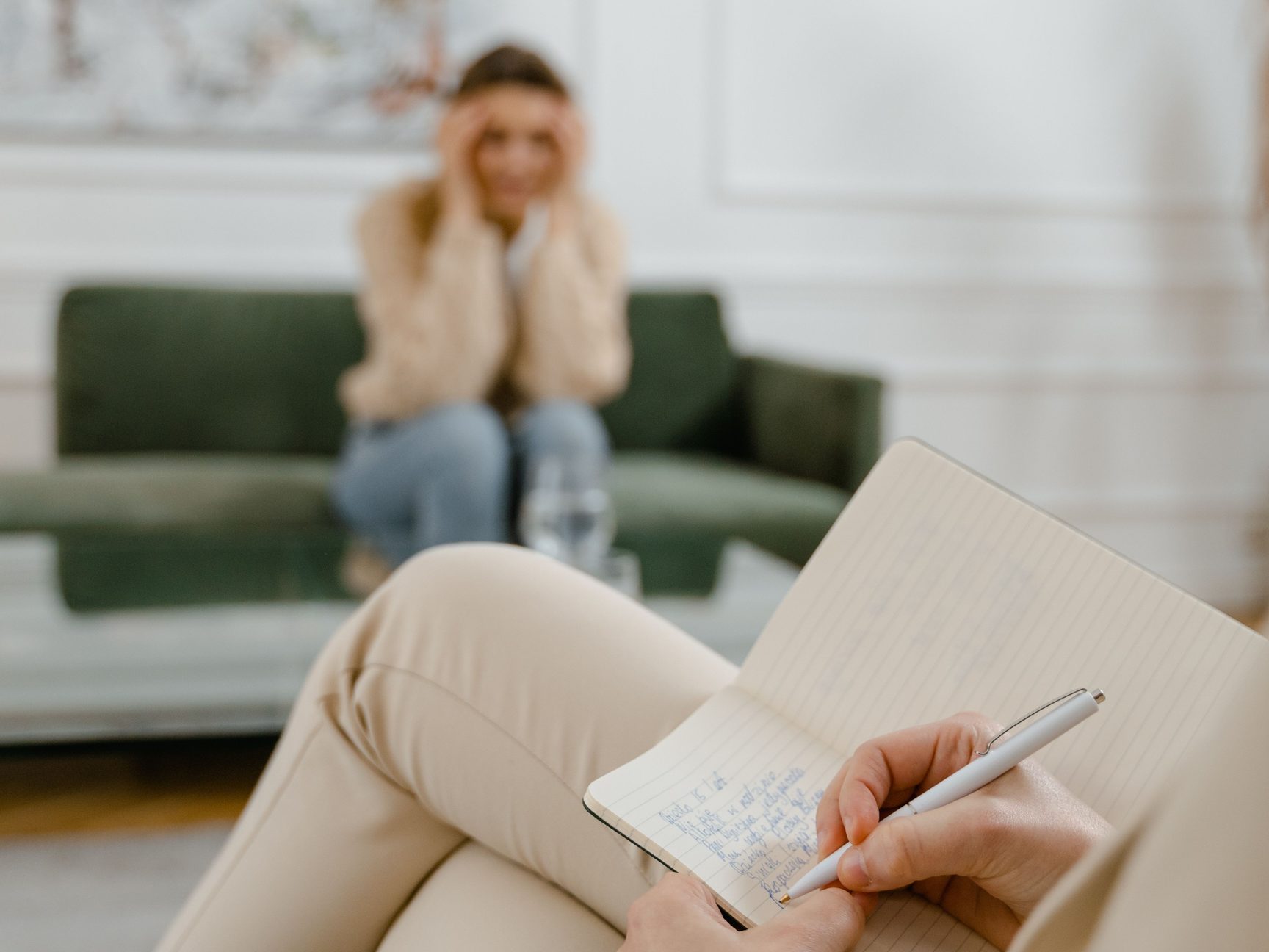A therapist’s hands writing in a notebook, with a client in soft focus on a green sofa across the room, her hands to her head