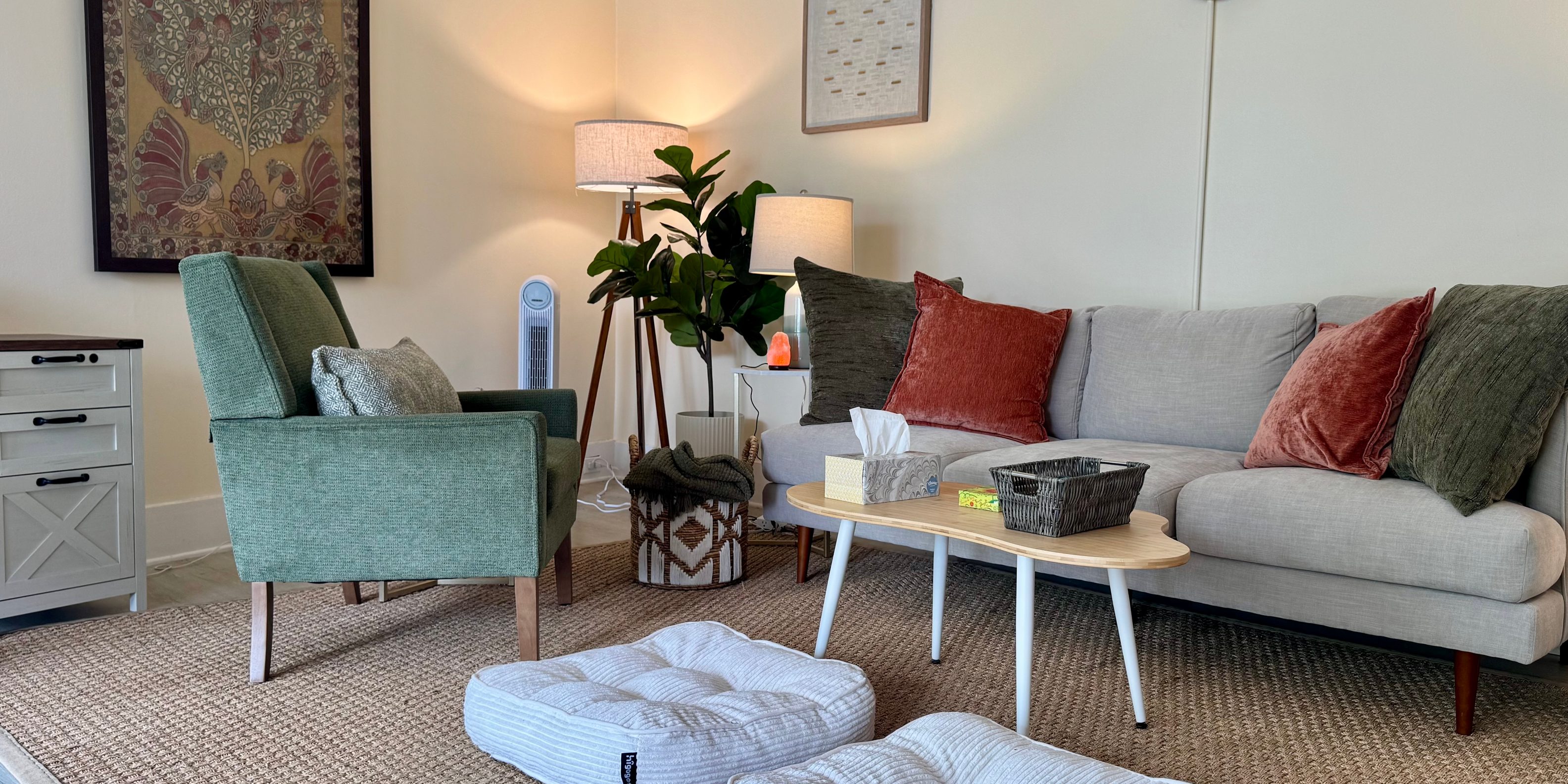 Tina's therapy room: a grey sofa with olive and rust cushions, a green armchair, floor cushions, and a framed Indian tapestry on the far wall