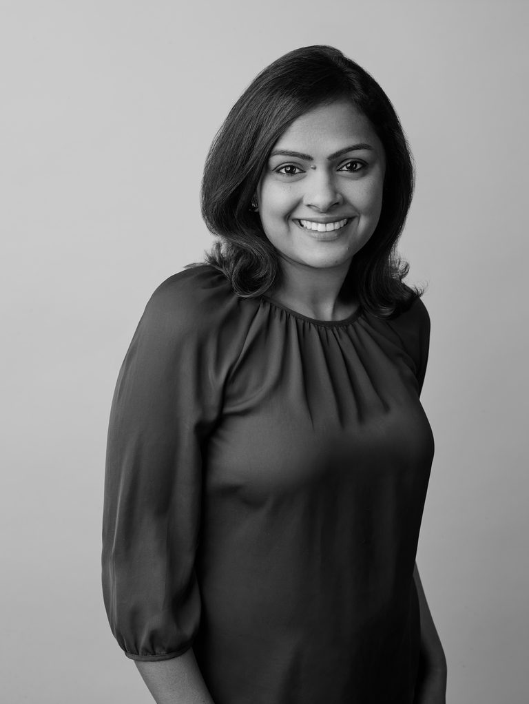 Black and white portrait of Tina Aggarwal smiling at the camera, in a dark blouse with a gathered neckline, against a plain grey background